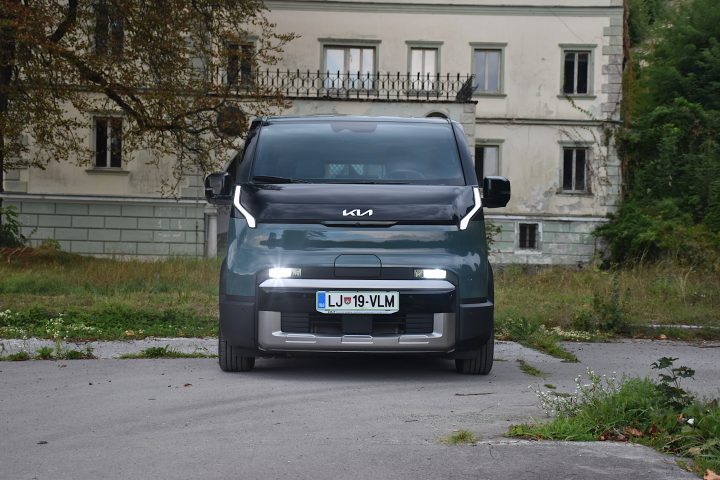 Temno zelena električna Kia PV5 Passenger, parkirana v naravnem okolju pred starejšo stavbo, z drevesi in zelenjem v ospredju