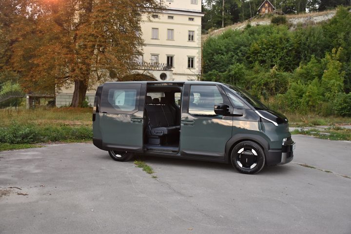 Temno zelena električna Kia PV5 Passenger, parkirana v naravnem okolju pred starejšo stavbo, z drevesi in zelenjem v ospredju - poudarek na zadnjih drsnih vratih