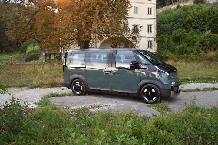 Temno zelena električna Kia PV5 Passenger, parkirana v naravnem okolju pred starejšo stavbo, z drevesi in zelenjem v ospredju