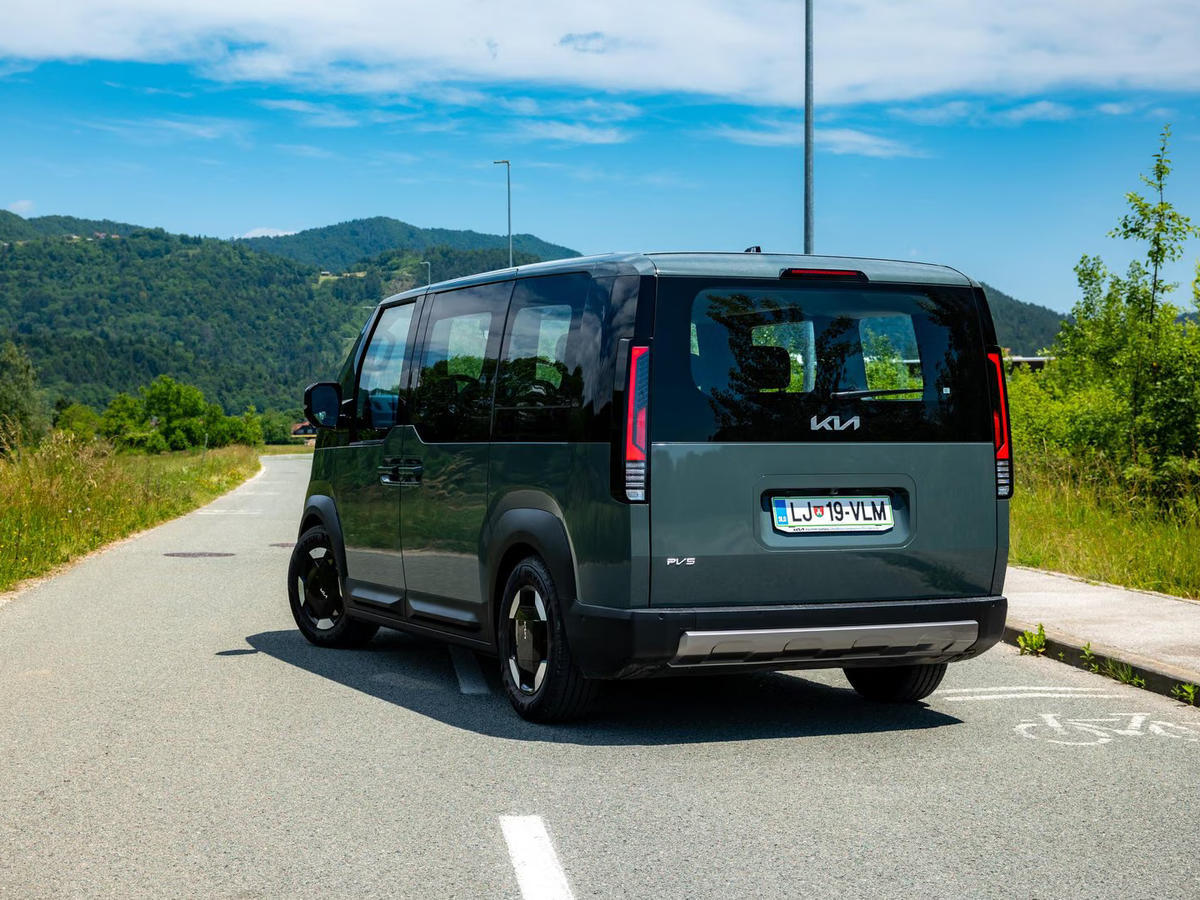 Električni Kia PV5 Passenger, pogled od zadaj na podeželski cesti z zelenimi griči v ozadju.